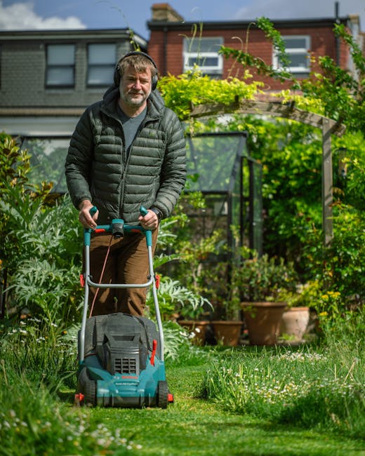 A middle-aged man with short, grey hair and a beard is operating a lawn mower in the garden of a residential property in Lewisham. He is wearing a grey puffer jacket, a dark shirt underneath, and brow